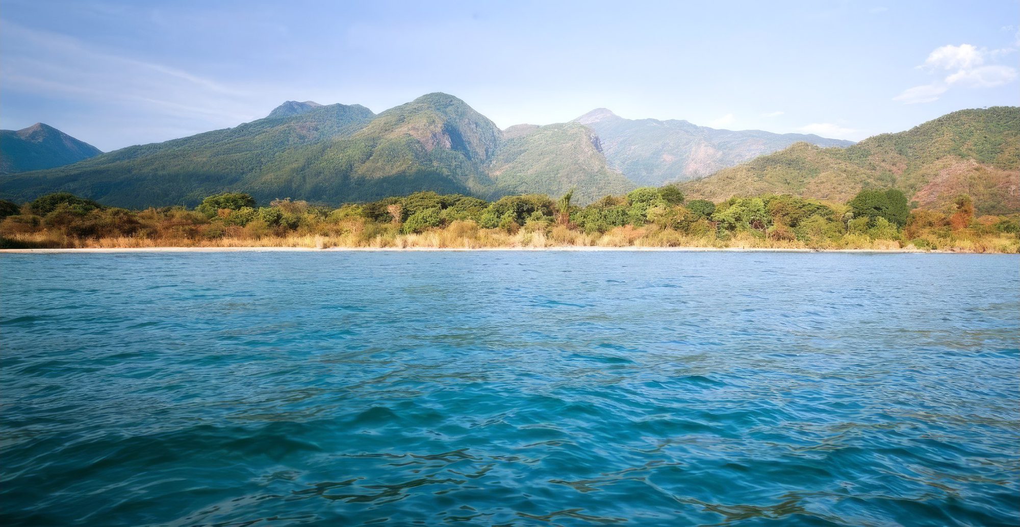 Agua turquesa con olas ante una orilla de arena y montañas boscosas en el Parque Nacional de Mahale Mountains